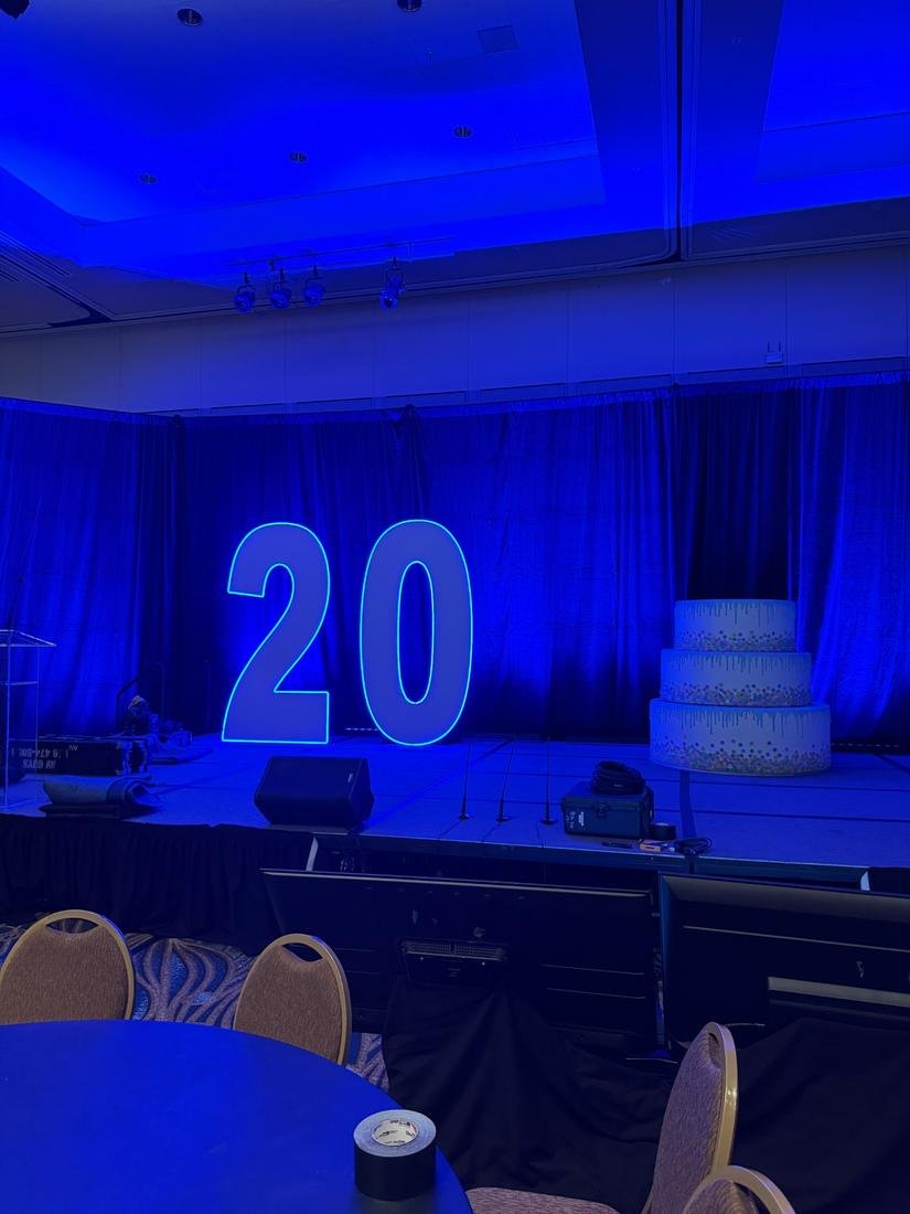 Giant marquee number 20 outlined in blue LED on a stage beside a large three tier cake prop