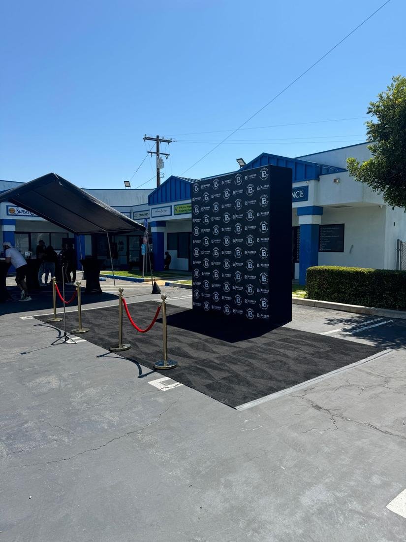 Black step and repeat wall with a repeating logo, black carpet and gold stanchions with red rope in a parking lot